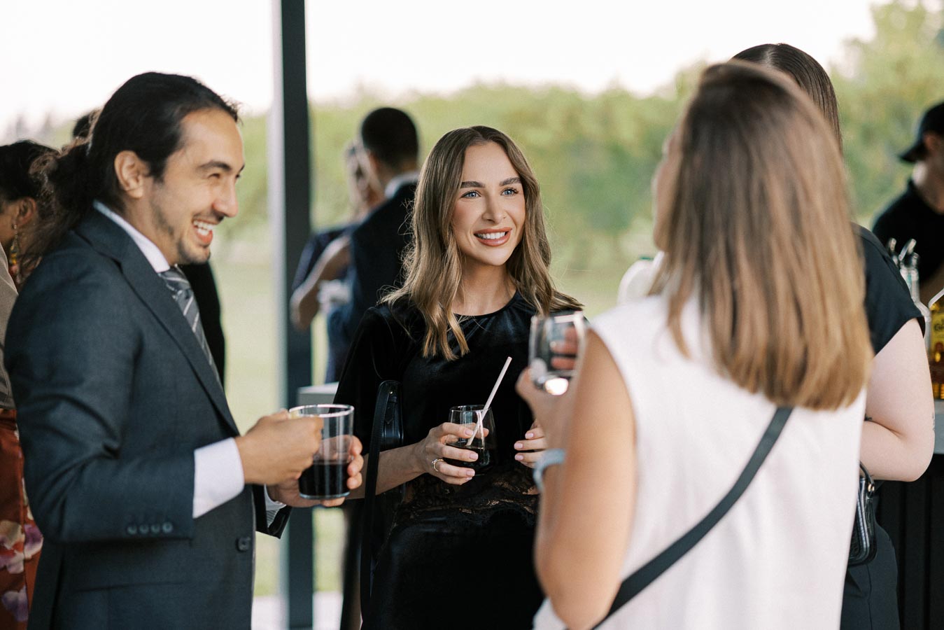 Group of people socializing at an outdoor event, dressed in formal attire, holding drinks and smiling.