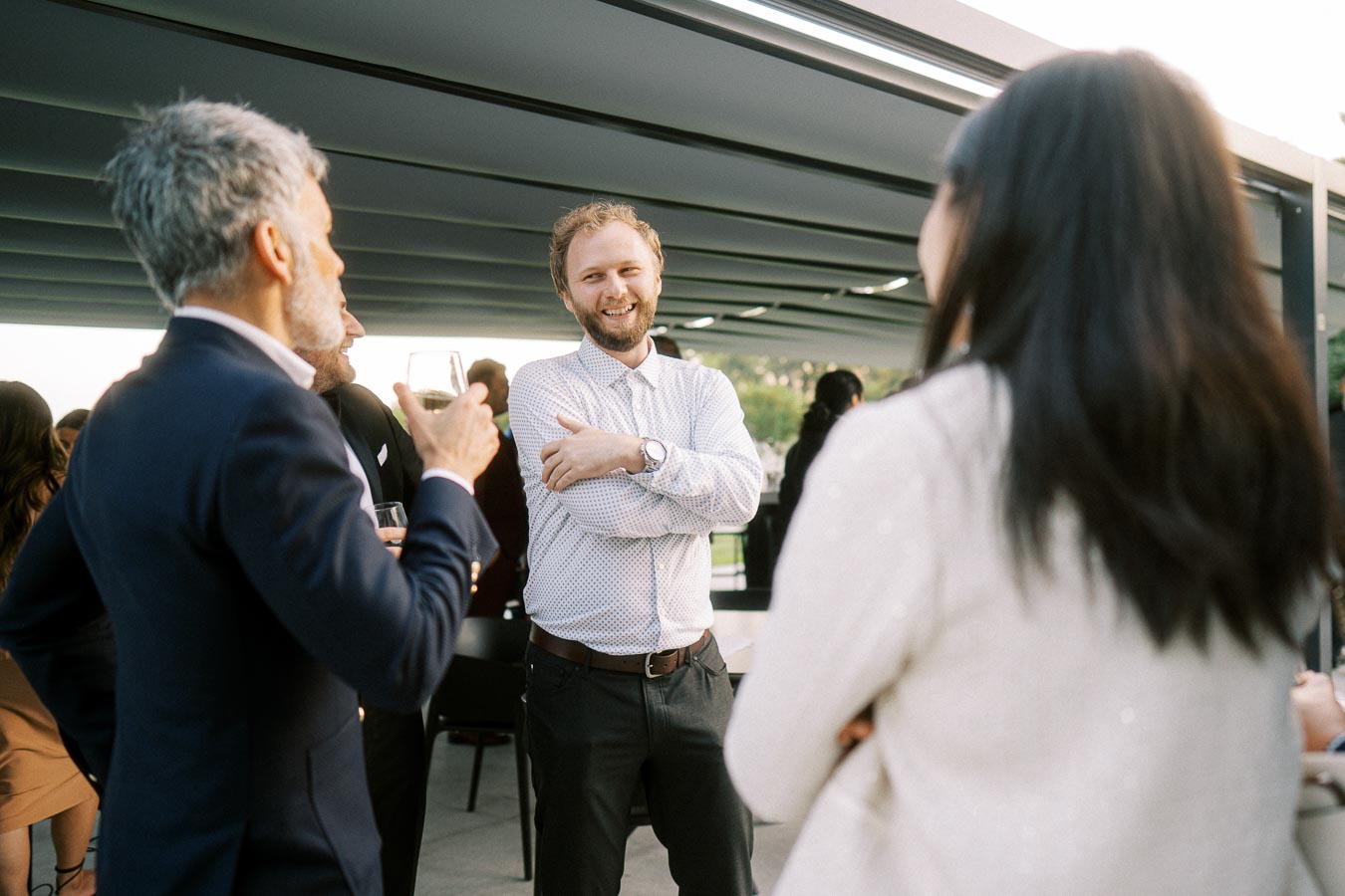 A group of people engaging in conversation at an outdoor social event, with one person in a striped shirt smiling and others holding drinks under a covered patio. The atmosphere is casual and friendly.