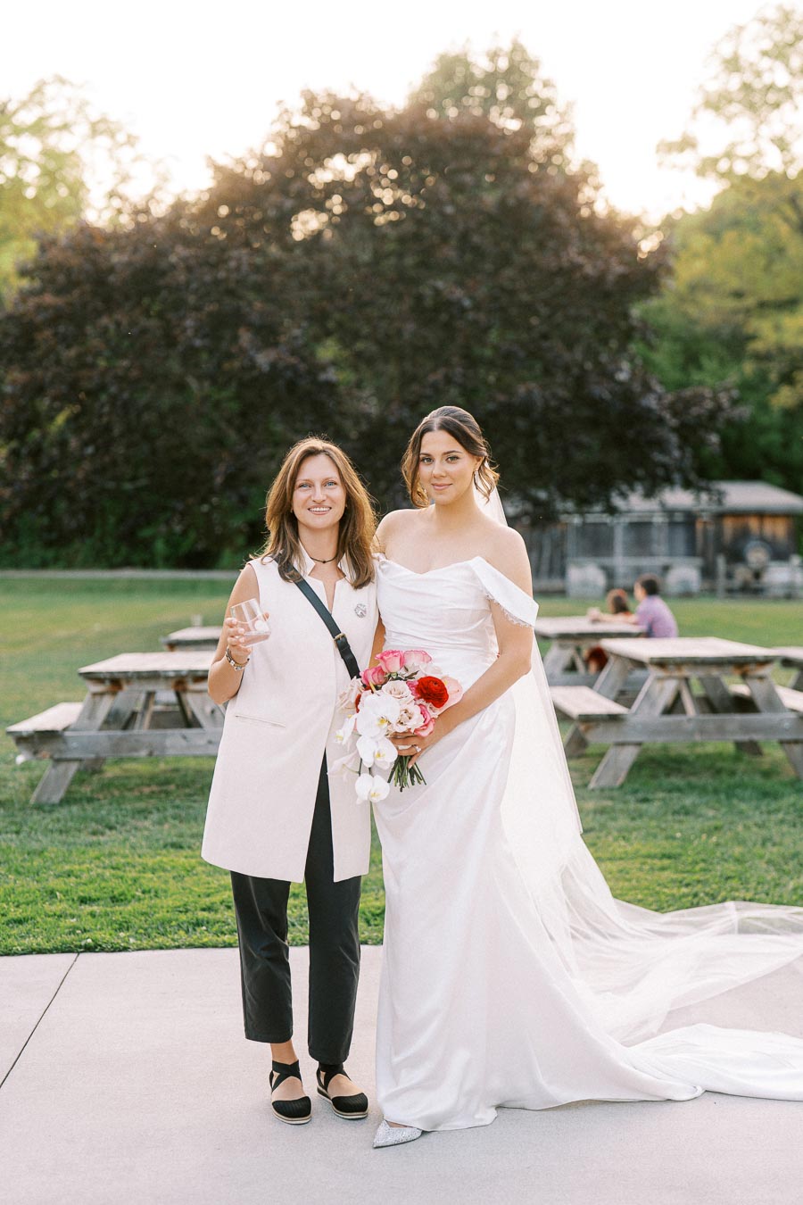 Bride in elegant white wedding dress holding a bouquet stands next to a smiling woman in a light vest, outdoors with picnic tables and lush greenery in the background.