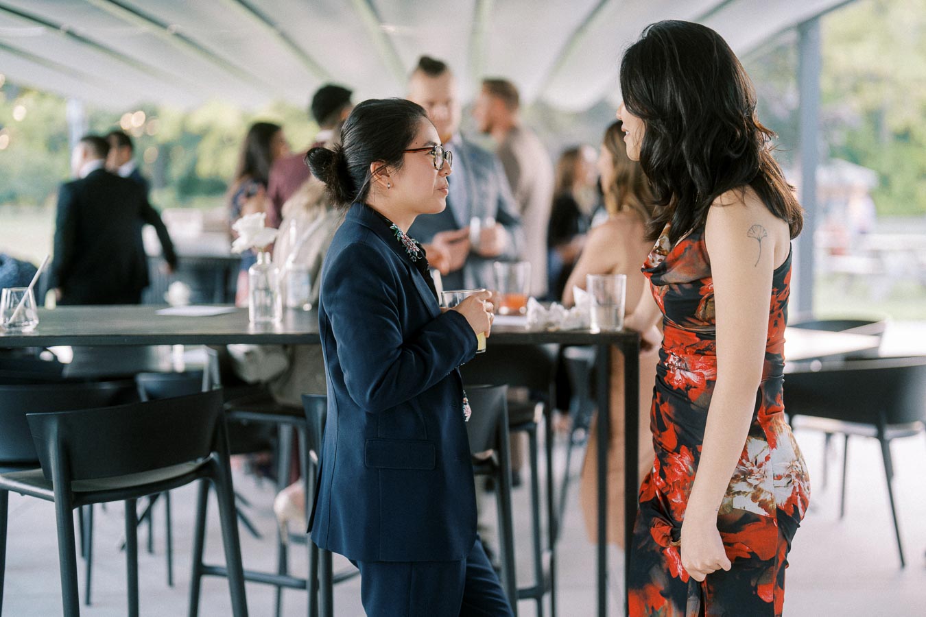 Two people having a conversation at a stylish outdoor event, one in a dark suit and the other in a vibrant floral dress, surrounded by a social gathering.