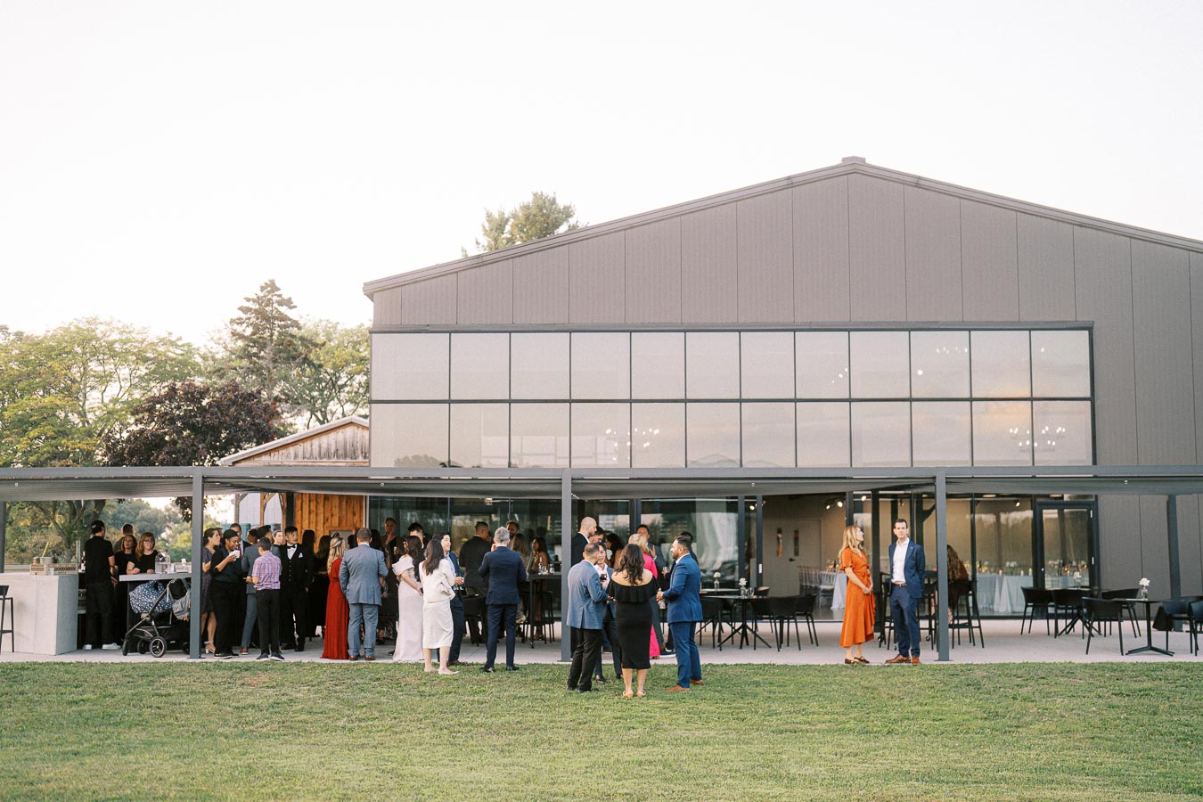 Alt text: Outdoor gathering of elegantly dressed guests at a modern venue with large windows, surrounded by green grass and trees, under a clear sky.