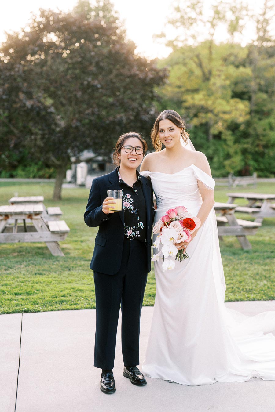 Two individuals celebrating outdoors; one in a white wedding dress holding a bouquet of flowers, and the other in a dark suit with a drink, standing on a lush green lawn with picnic tables and trees in the background.
