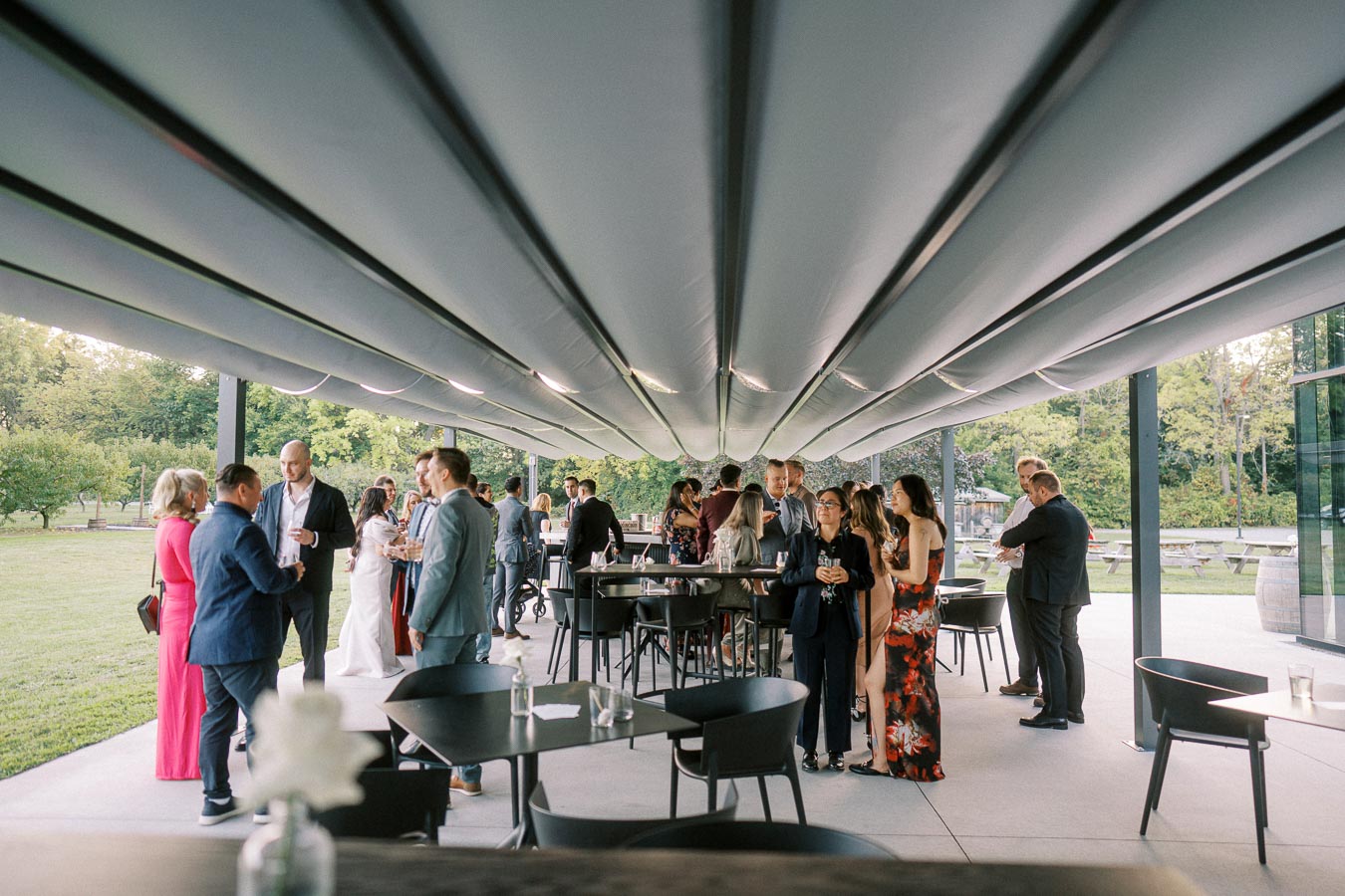 A group of people socializing at an outdoor event under a modern canopy, with a scenic view of greenery and trees in the background.