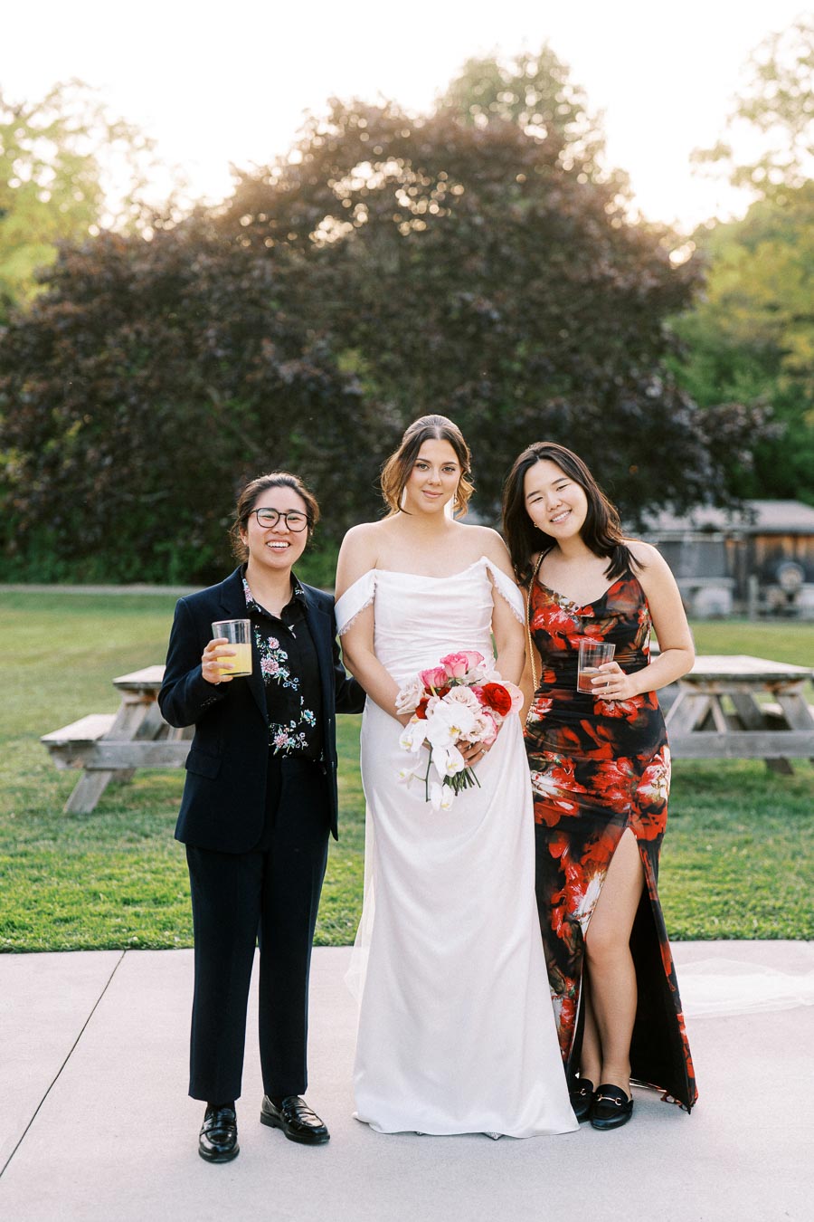 Three people posing together at an outdoor wedding, with one person in a white wedding dress holding a bouquet of flowers and the others in formal attire, standing on a grassy area with trees in the background.