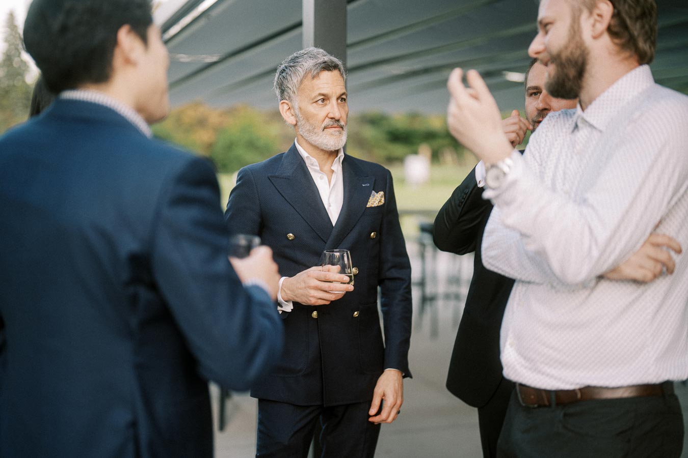 Group of men in formal wear engaging in conversation at an outdoor event, one holding a glass.