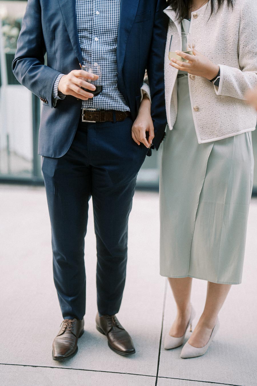 Stylish couple attending an outdoor event, with the man in a blue suit holding a drink, and the woman wearing a white blazer and dress, showcasing elegant fashion.