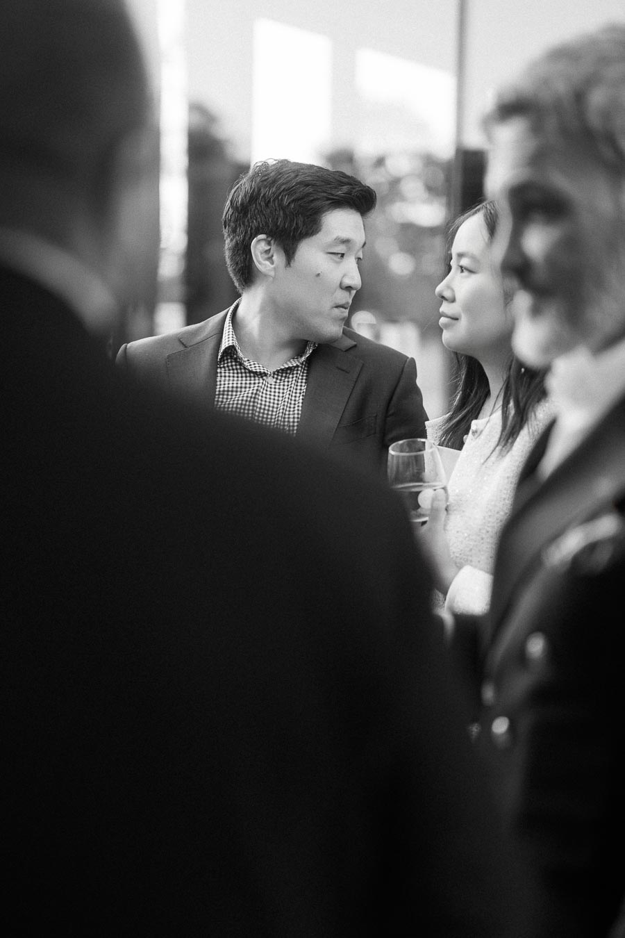 A black-and-white photo capturing a formal social event, featuring a man and woman in conversation amidst a group of people. The man is wearing a suit, and the woman is holding a wine glass, suggesting a networking or corporate gathering setting.