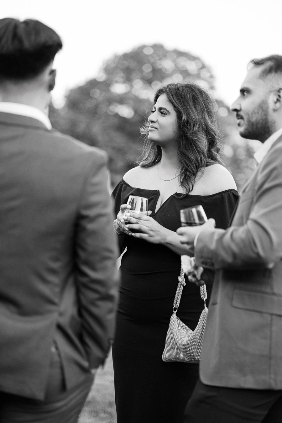 Black and white photo of three elegantly dressed individuals at an outdoor social event, holding wine glasses and engaged in conversation.