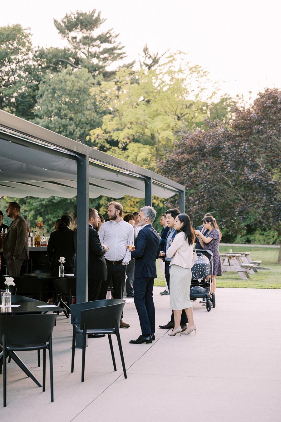 Outdoor gathering of people in formal attire standing near a modern patio bar, set against a backdrop of lush green trees and a picnic area.