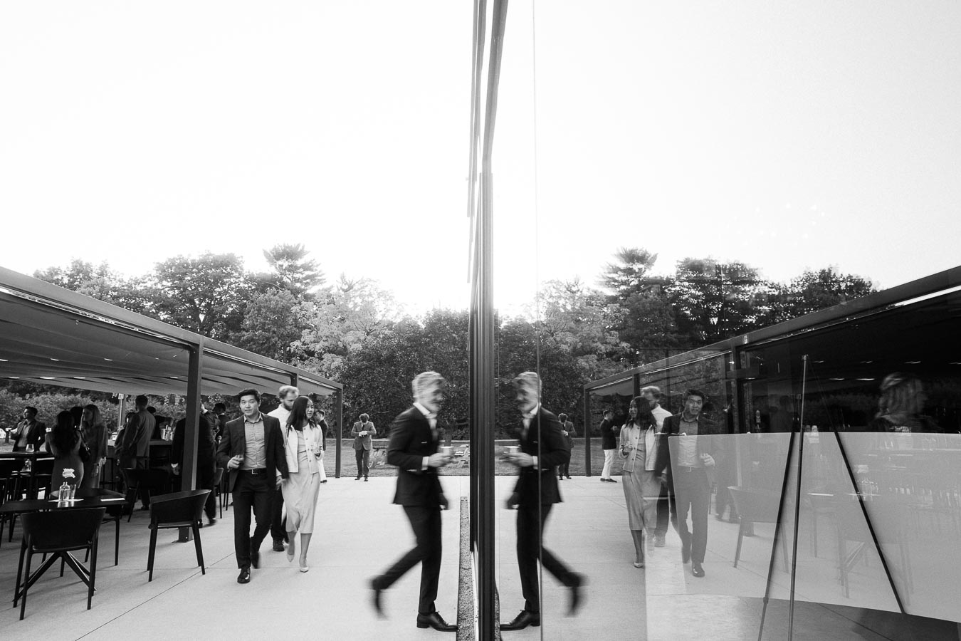 Black and white image of people walking outside a modern glass building during an outdoor event. The scene captures reflections in the glass, with guests dressed in formal attire, mingling under a canopy surrounded by trees.
