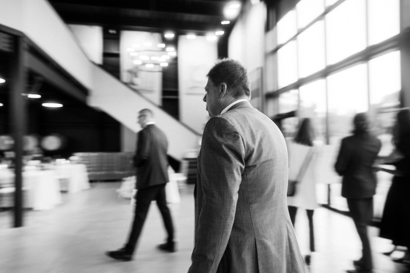 Blurred black and white photo of several people in formal wear walking through a modern, well-lit lobby with large windows and a staircase in the background.