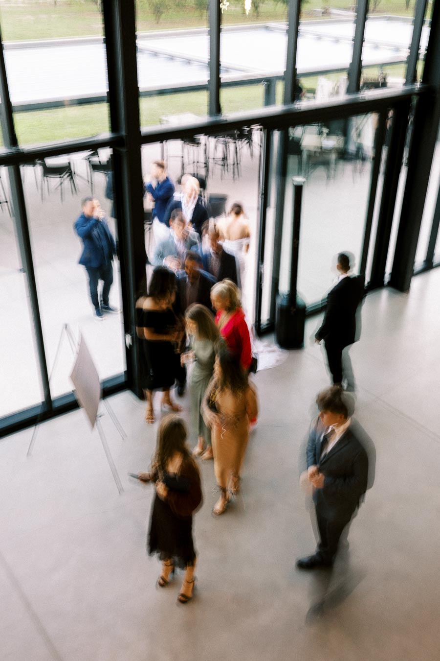 A group of elegantly dressed people socializing in a modern glass-walled event space, with some individuals standing near the entrance and others mingling indoors, creating a lively and sophisticated atmosphere.