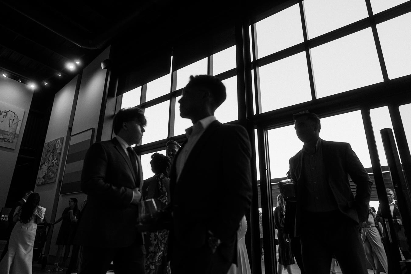 Black and white image of a formal gathering with people in suits and dresses socializing near large windows.