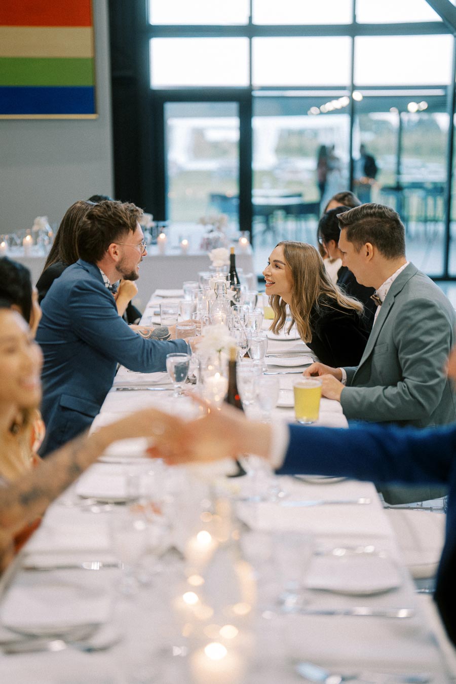 Group of elegantly dressed people enjoying a lively conversation at a long dining table, adorned with candles and wine glasses, in a modern, bright restaurant setting with large windows.
