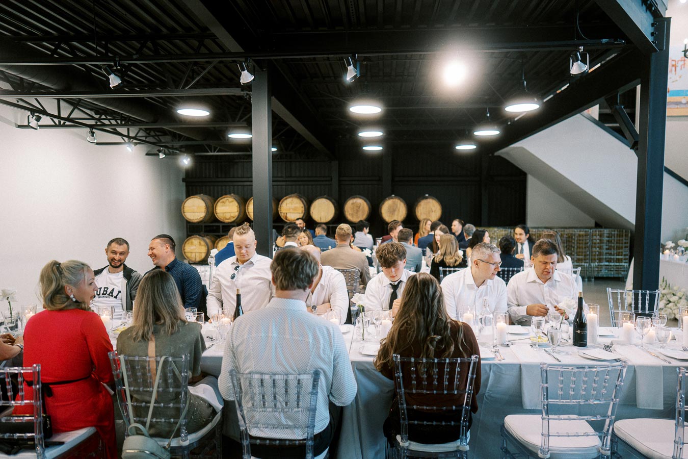 A group of people enjoying a dinner event in a modern winery setting, featuring wine barrels as decor. The attendees, dressed in smart casual attire, are seated at a long table with white tablecloths, surrounded by ambient lighting that enhances the festive atmosphere.