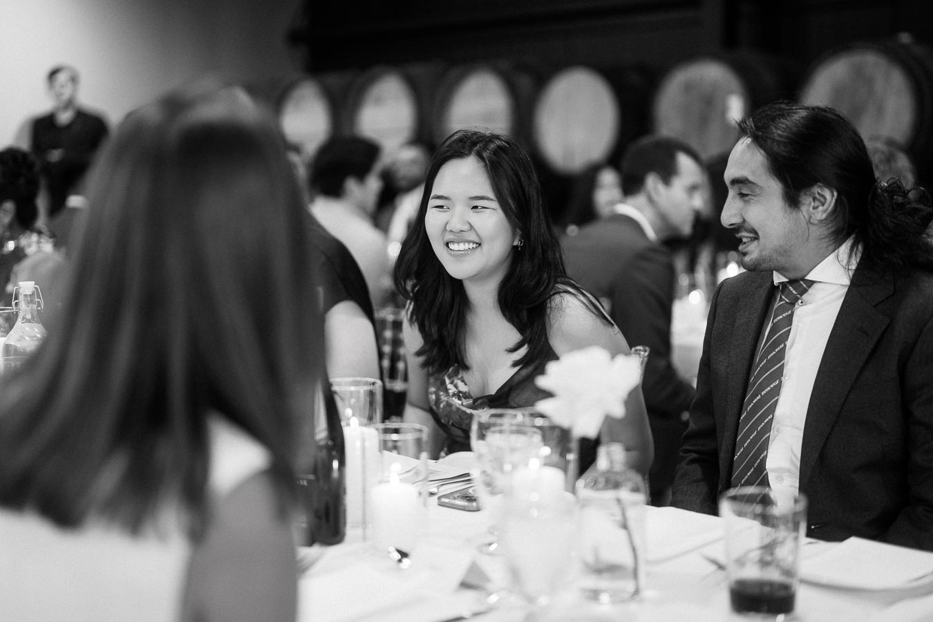 Black and white photo of a smiling woman at a formal event, seated at a table with glasses and candles, engaging in conversation with a man in a suit. In the background, blurred wine barrels and other guests are visible, creating a warm and lively atmosphere.