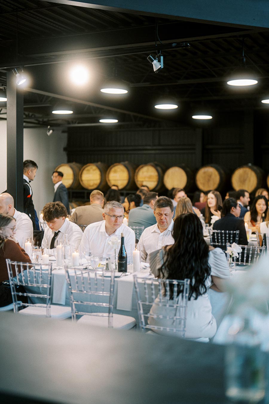 Guests seated at elegantly decorated tables during a wedding reception in a winery-themed venue with barrels in the background, ambient lighting creating a warm atmosphere.