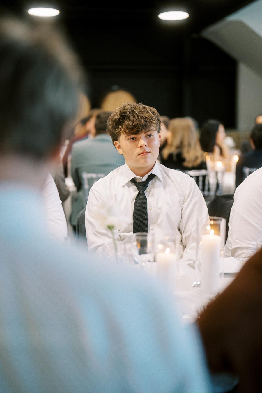 A young man in a white shirt and black tie sitting at a formal event, surrounded by people and lit candles, creating an elegant atmosphere.