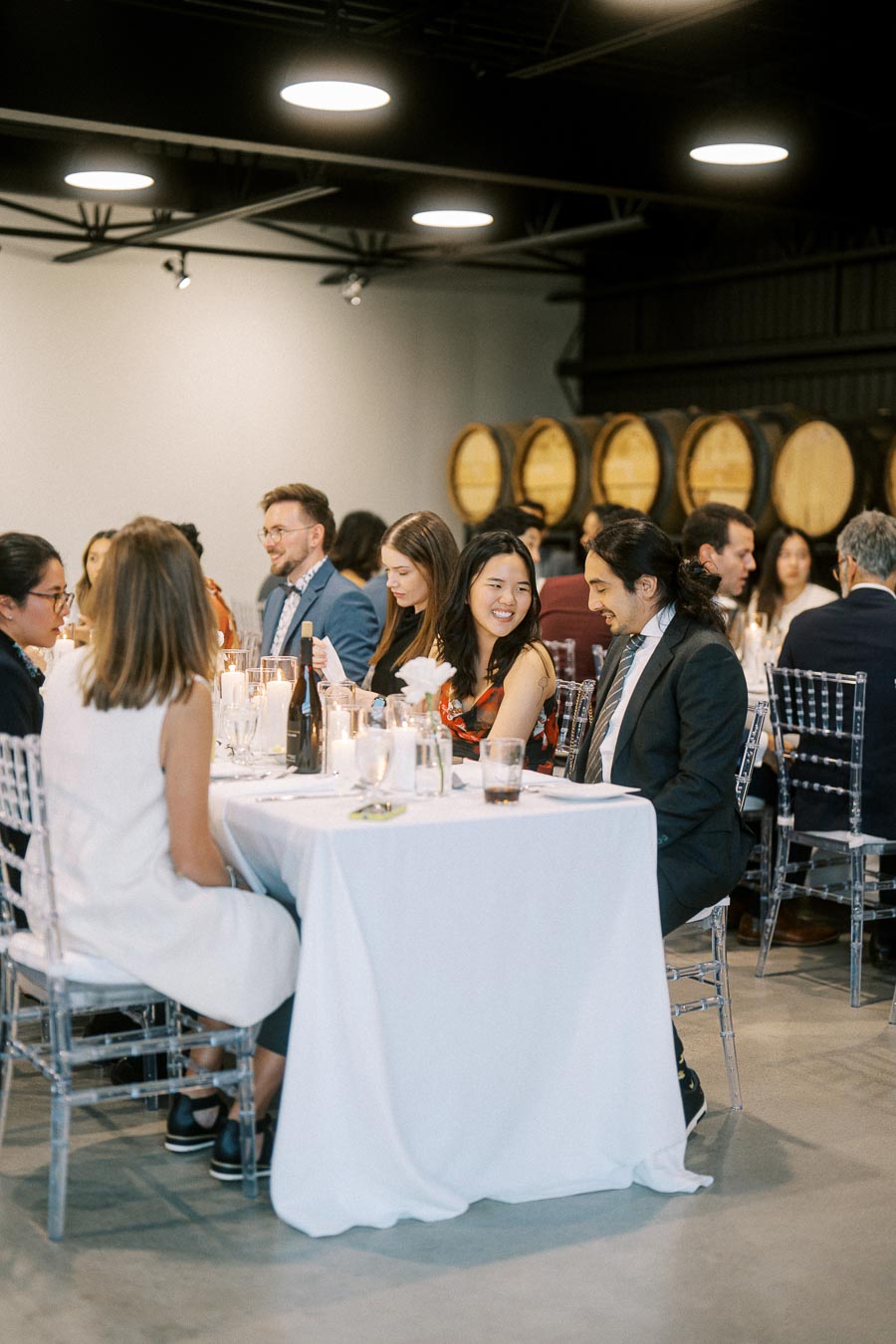 A group of people in formal attire enjoying a dinner event in a stylish venue with wooden barrels in the background, creating an elegant atmosphere.