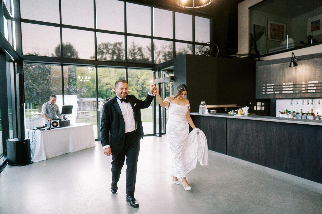 Bride and groom joyfully entering a modern reception venue, with a DJ in the background, during their wedding celebration.