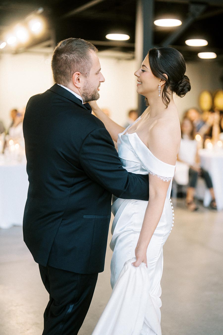 A bride and groom sharing a dance at their wedding reception, with the bride in a white off-shoulder gown and the groom in a black suit, surrounded by softly lit candles and guests in a modern venue.