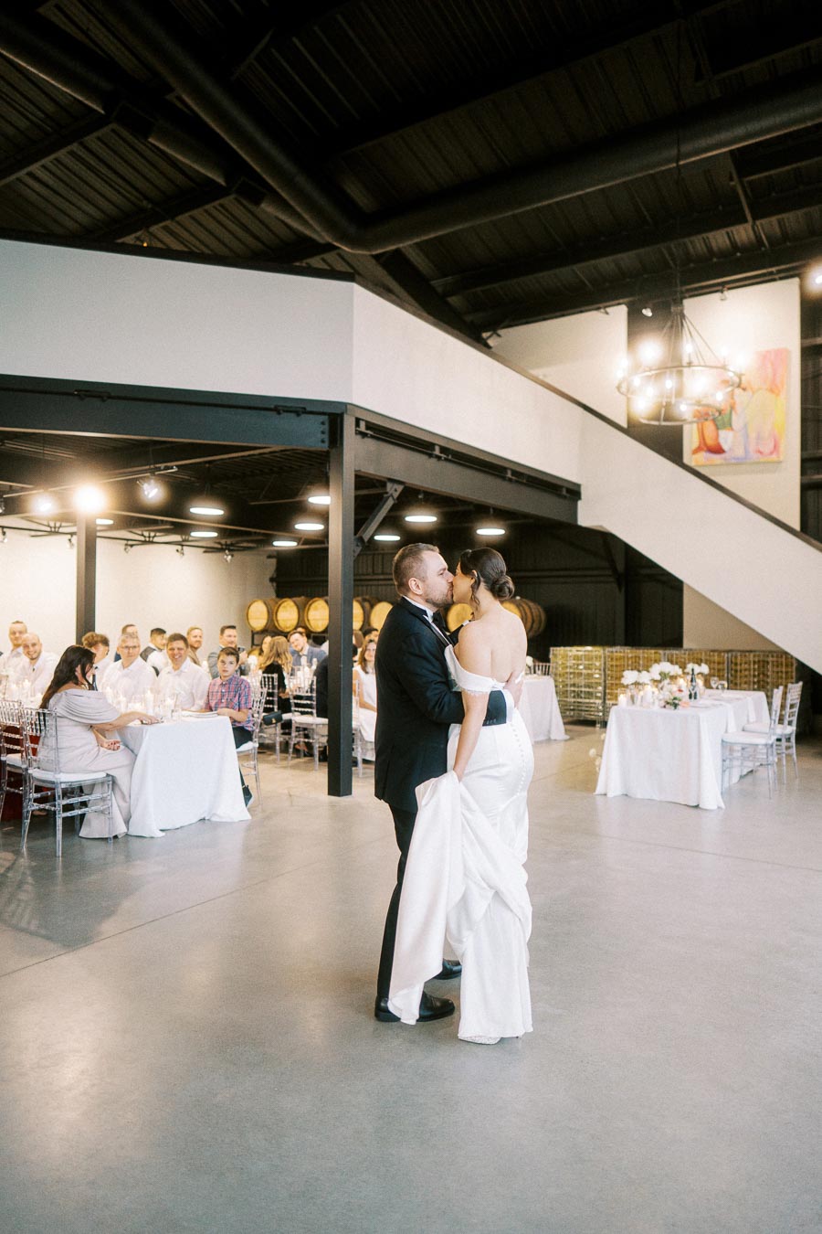 A bride and groom share a romantic first dance in a modern, elegantly decorated reception hall, with seated guests observing the moment.