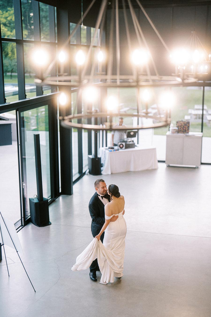 A bride and groom share a romantic first dance in a modern venue with large windows and elegant lighting fixtures, while a DJ plays music in the background.
