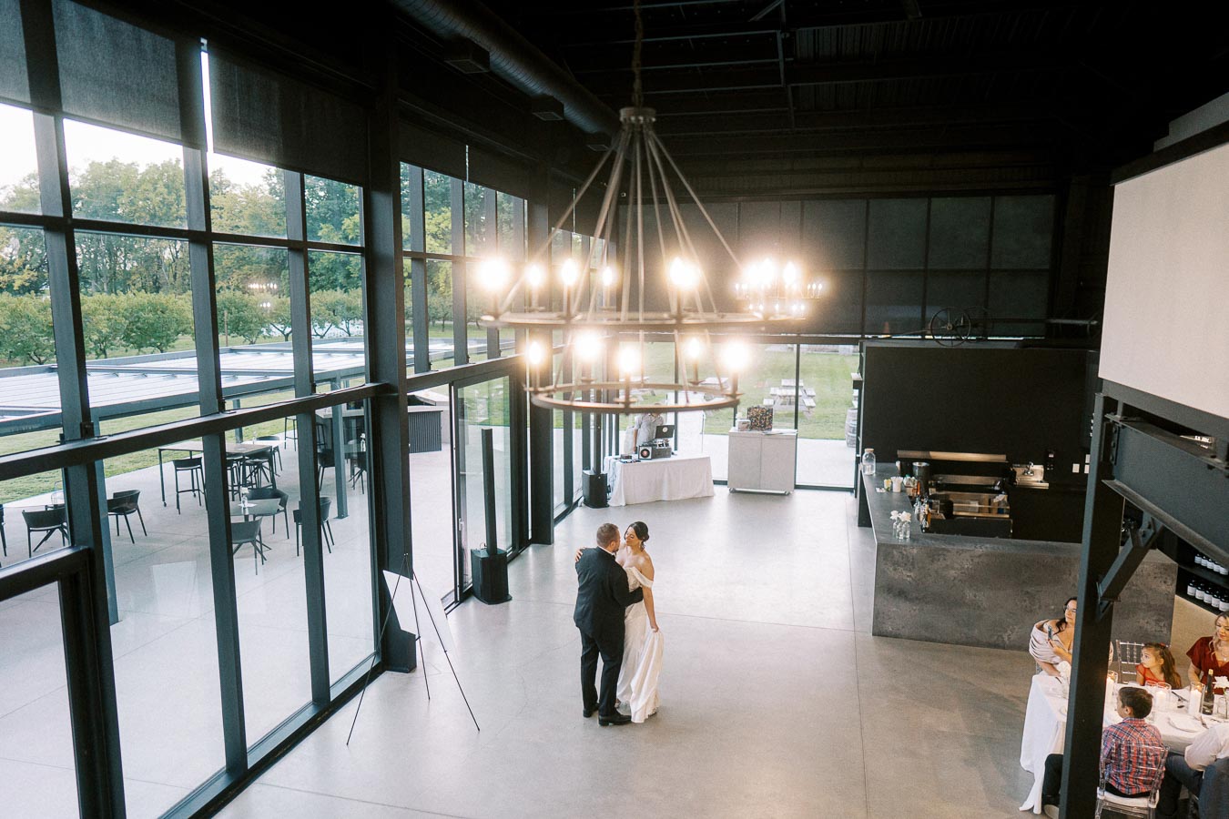 A couple shares a romantic dance in a spacious, modern venue with large windows and soft lighting, as guests seated at tables enjoy the ambiance.