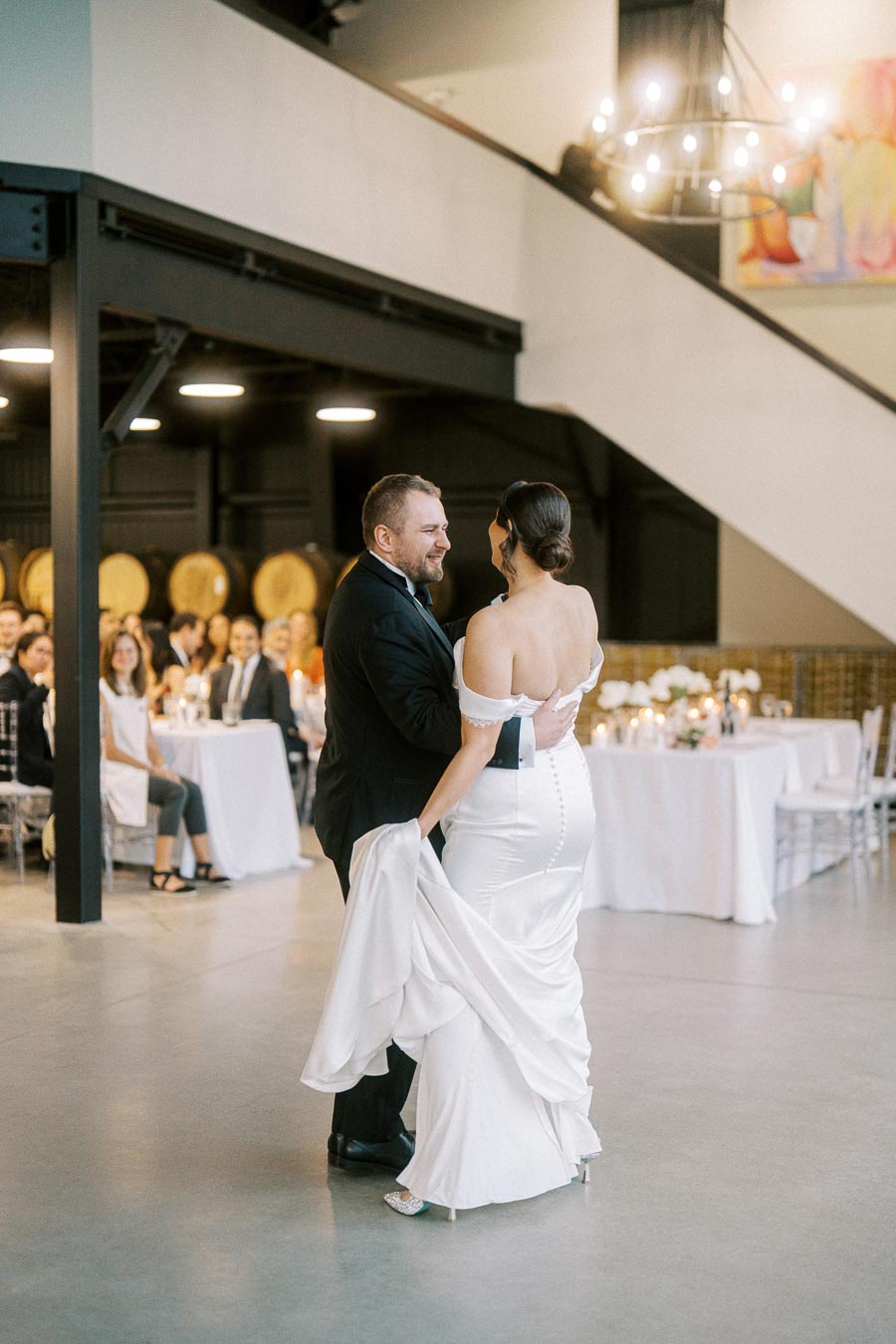 A couple shares a joyful first dance at their wedding reception in an elegant venue with guests seated at tables in the background, under a warm chandelier light.