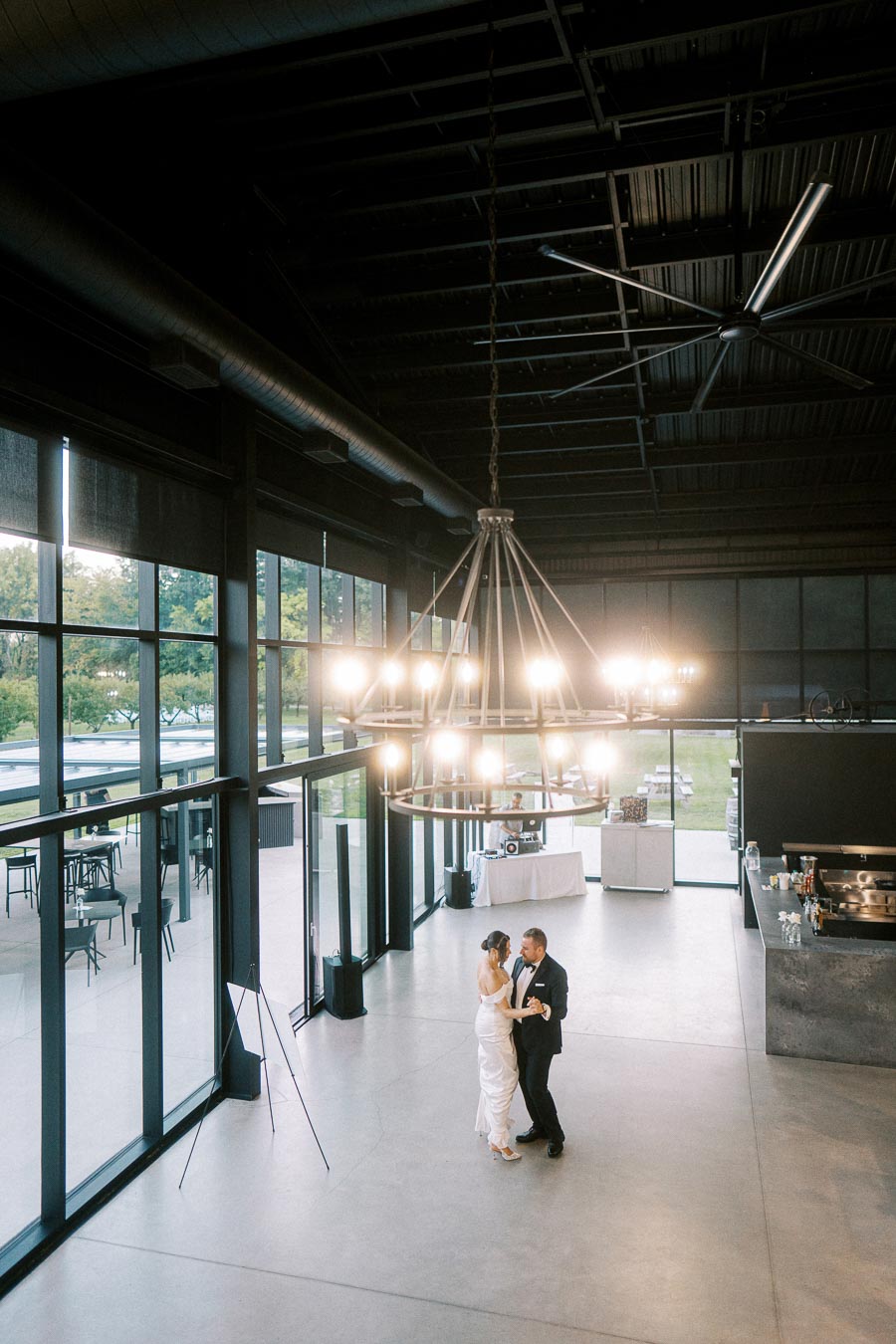 Elegant wedding couple dancing in a modern industrial venue with large windows and a grand chandelier overhead.