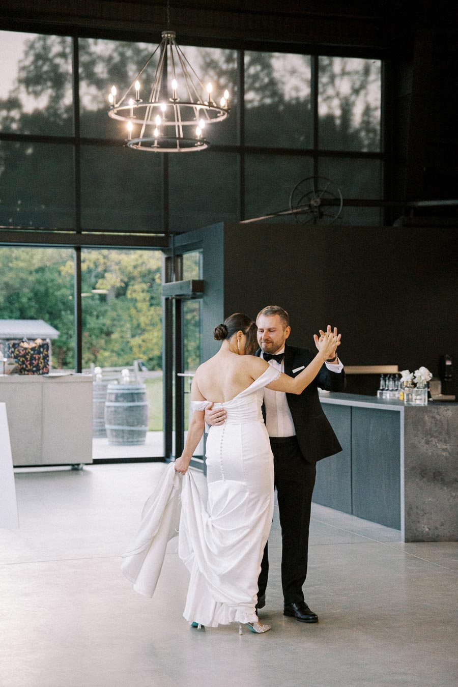 A couple elegantly dancing under a chandelier in a modern wedding venue with floor-to-ceiling windows, capturing a romantic moment.