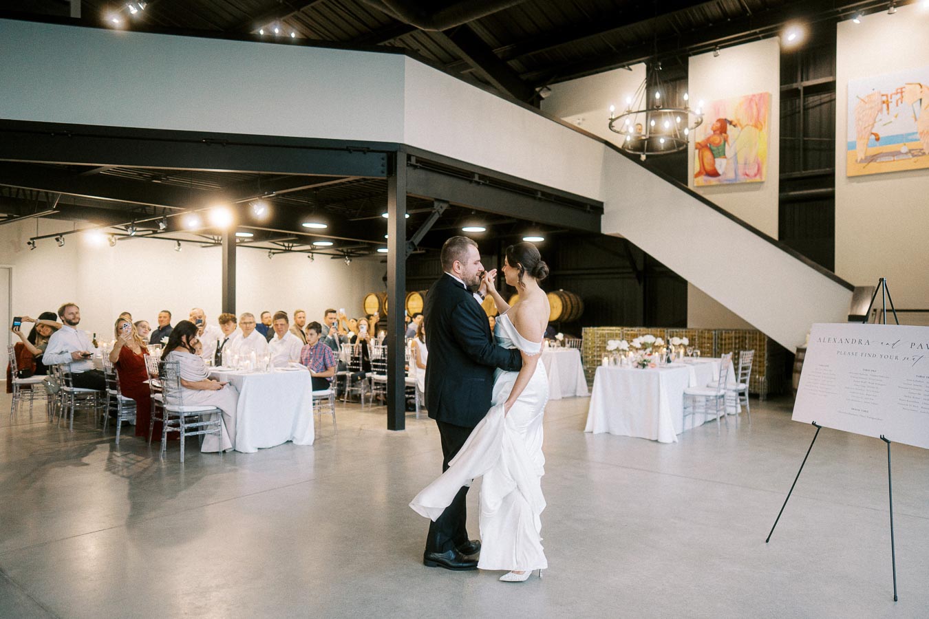 A bride and groom share their first dance at a wedding reception, surrounded by seated guests in a modern venue with high ceilings and contemporary art.
