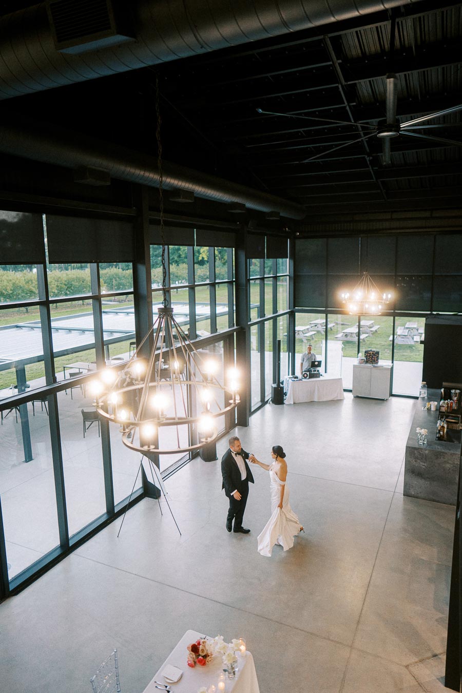 A bride and groom share a first dance in a modern, spacious venue with large windows overlooking greenery, featuring stylish chandeliers and elegant decor.