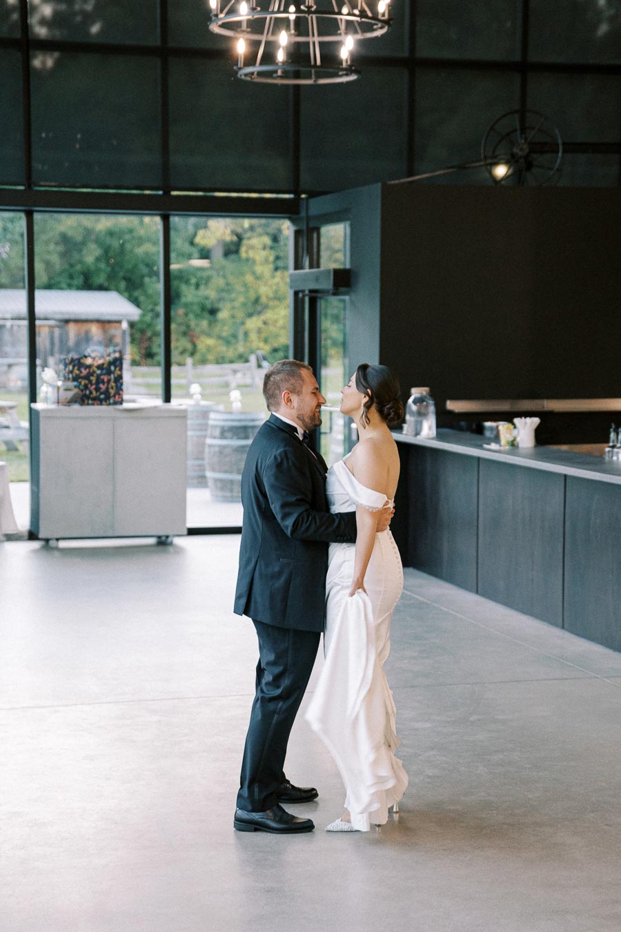 A couple sharing a romantic dance in an elegant indoor venue with large windows, featuring a modern chandelier and a scenic view of greenery outside. The bride is in an off-shoulder white gown and the groom is in a classic black tuxedo.