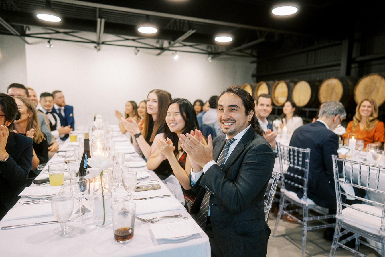 Guests clapping and smiling at a formal dinner event, seated at elegantly set tables with white tablecloths and floral centerpieces in a sophisticated venue.