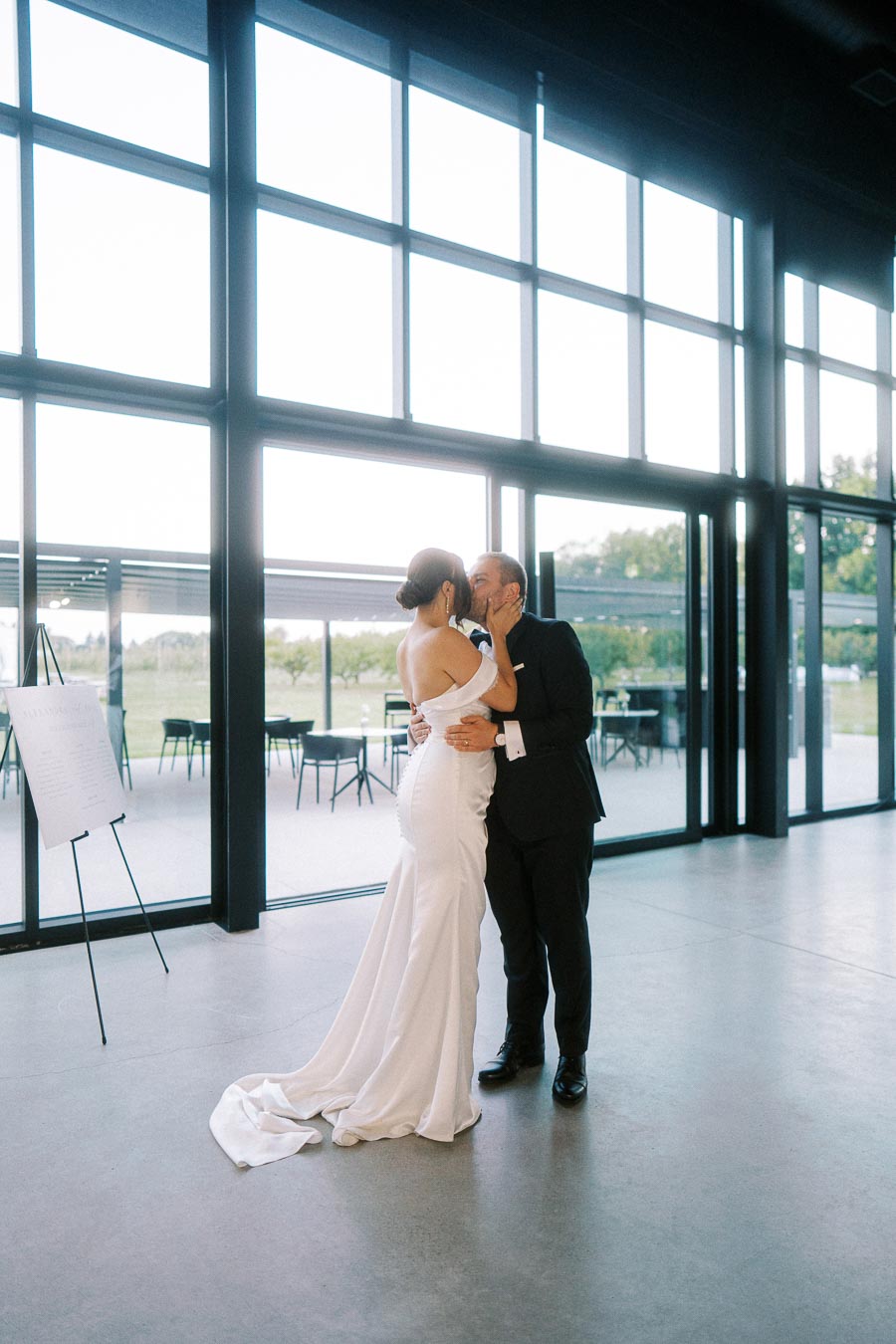 Bride and groom sharing a romantic kiss inside a modern, light-filled venue with large windows and an outdoor view.