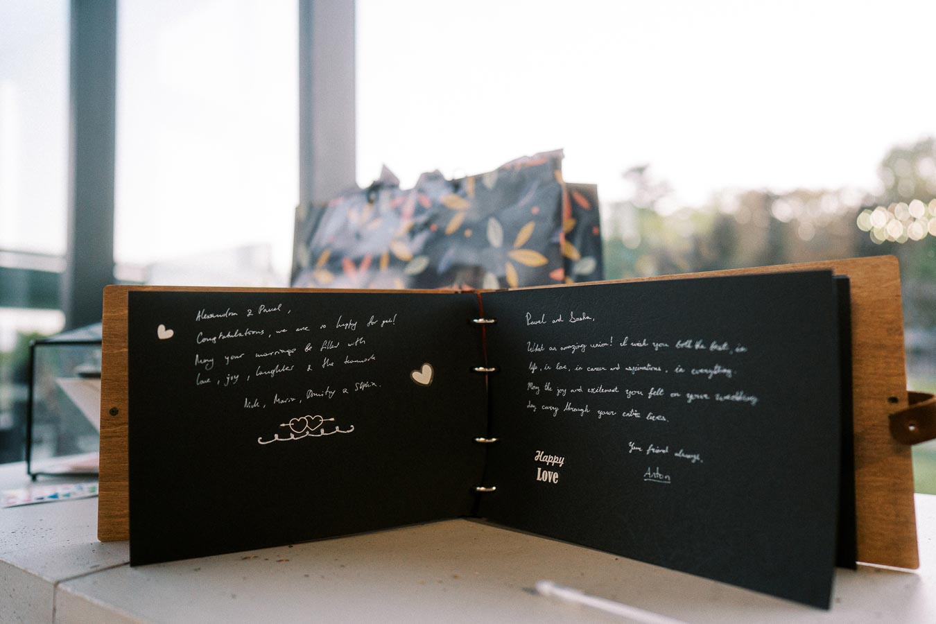Open wedding guestbook with handwritten messages on black pages, displayed on a light background, featuring heartfelt wishes and love-themed decorations.