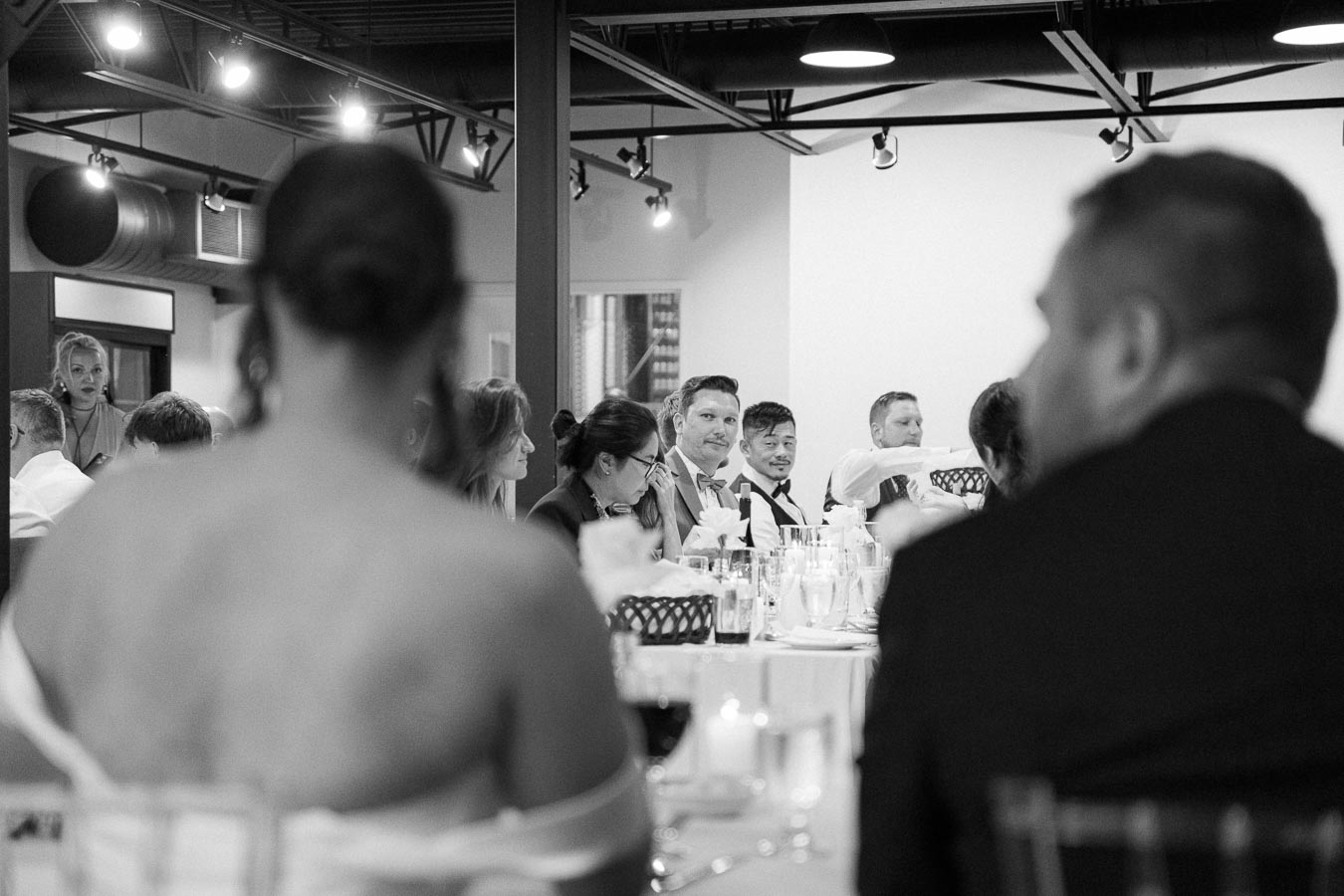 Black and white photo of people seated at a formal event, with tables elegantly set with wine glasses and centerpieces, in a modern venue with overhead lighting.