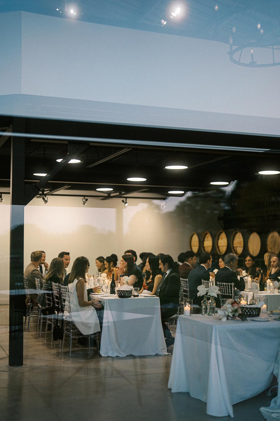 A group of people enjoying a formal dining event in a modern, warmly-lit venue with white tablecloths and decorative candles, viewed through a large window.