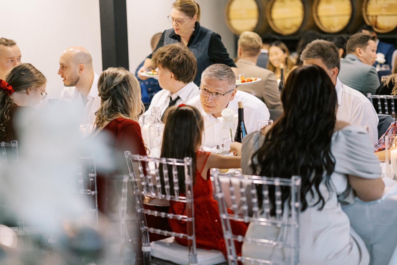 A diverse group of people at a formal dinner event, seated around a table with white linens and decorated with wine bottles and flowers. The setting appears to be a winery or banquet hall with barrels visible in the background.