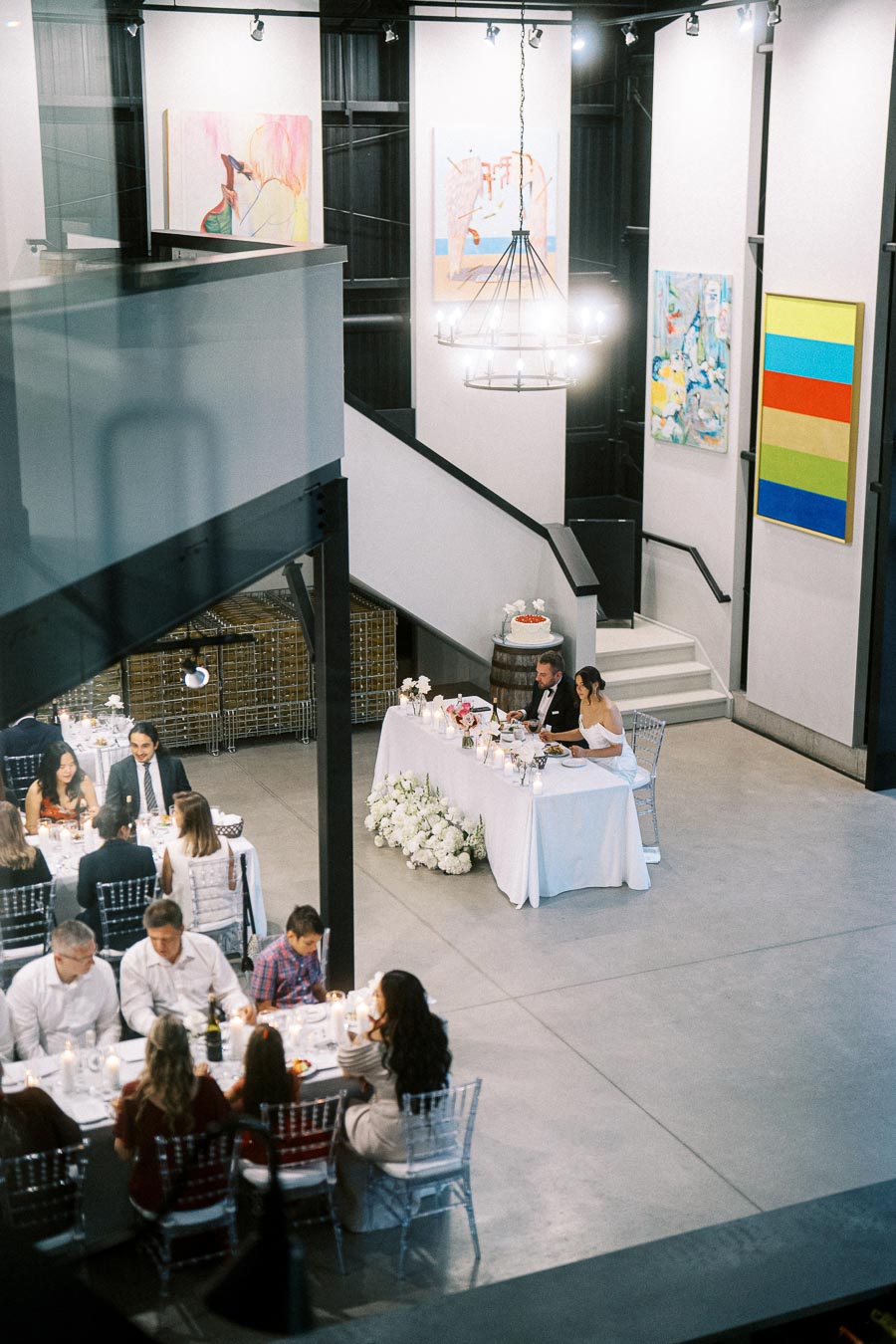 A wedding reception in a modern art gallery setting featuring elegantly dressed guests seated at tables, a couple at a decorated sweetheart table, abstract artwork on the walls, and a cake on display near the staircase.