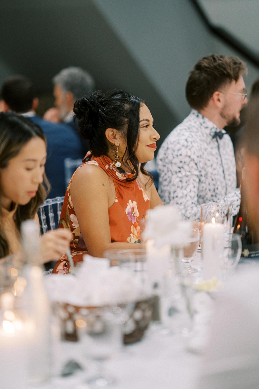 Elegant woman in floral dress attending a candlelit dinner event, surrounded by friends at a beautifully set table with glassware and soft lighting.