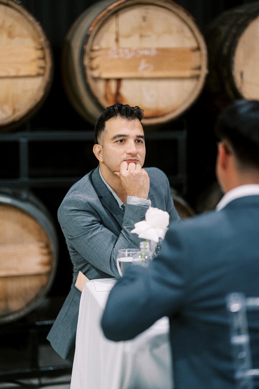 Man in a suit sits thoughtfully at a table in a winery, with wooden barrels in the background, during a business meeting or event.