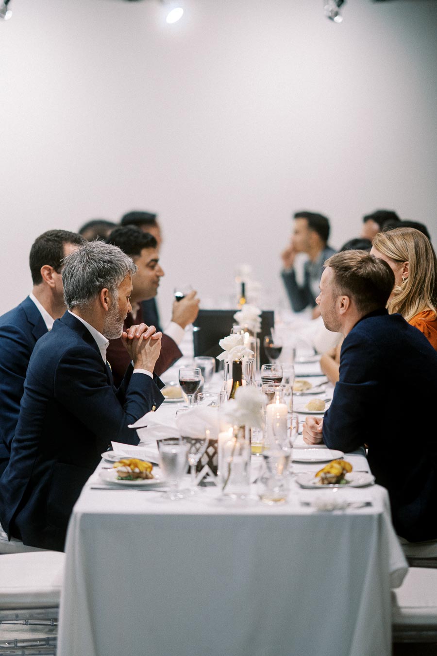 Group of people in formal attire engaged in conversation at an elegant dinner table setting with white tablecloths, candles, and fine dining items.