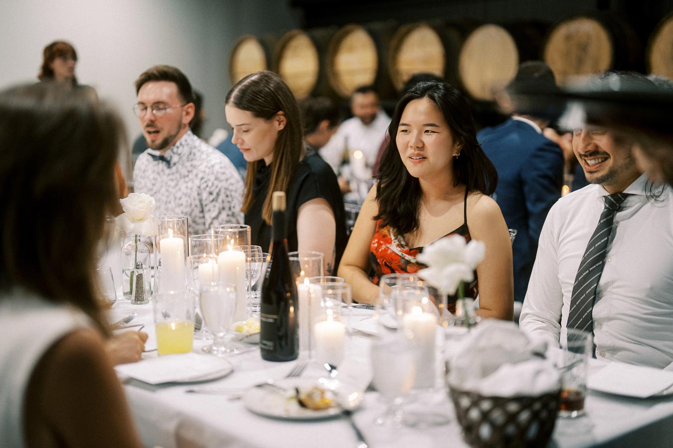A group of people enjoying a candlelit dinner at a sophisticated event, with wine and decorations, in a room with wooden barrel decor.