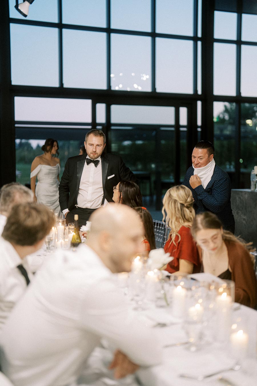 A group of people dressed in formal attire gather around a candle-lit dinner table at an elegant indoor wedding reception. The large windows in the background show a view of the evening sky.