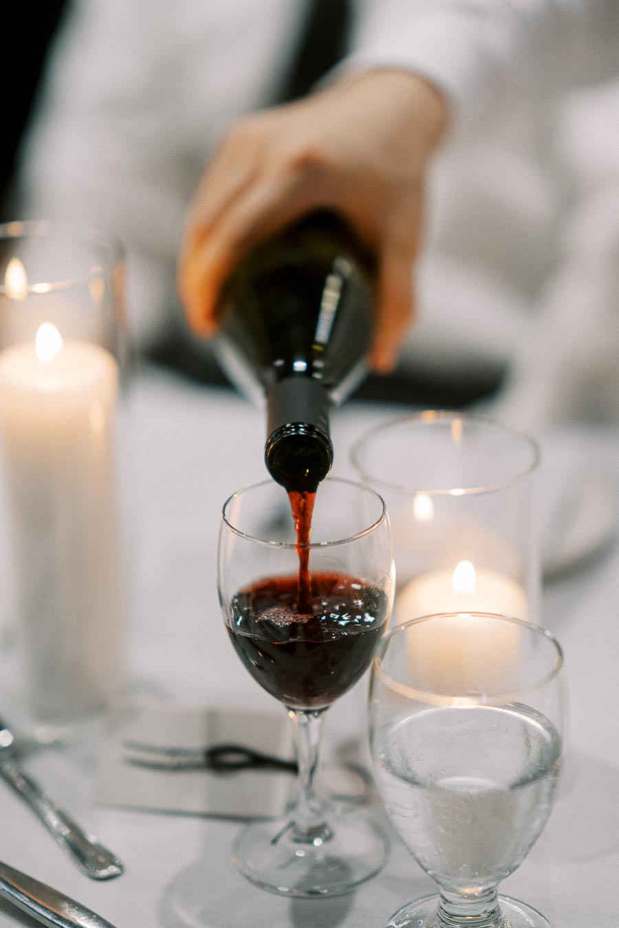 A hand pouring red wine into a glass on an elegantly set dinner table, accompanied by lit candles and glassware, creating a romantic and sophisticated ambiance.