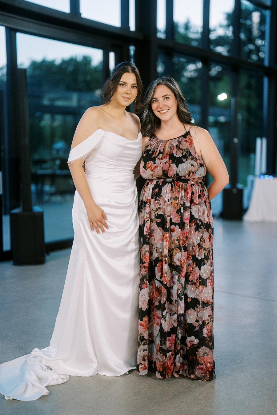 Two women posing together indoors; one wearing an elegant white wedding dress, the other in a floral print dress, with a modern glass window backdrop.