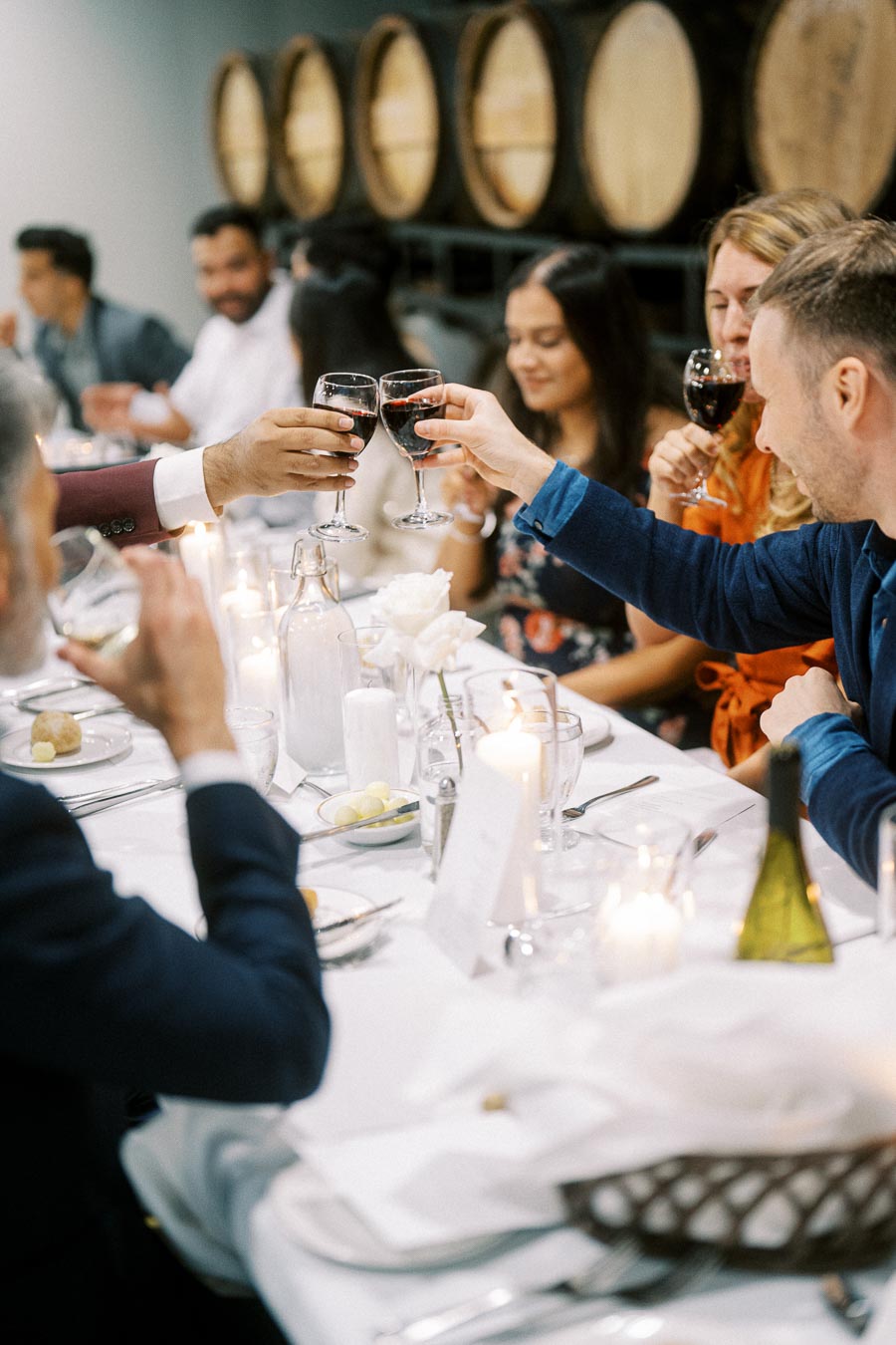 A group of people enjoying a wine toast at a banquet table in a winery, with candles and wine barrels in the background, creating a warm and festive atmosphere.