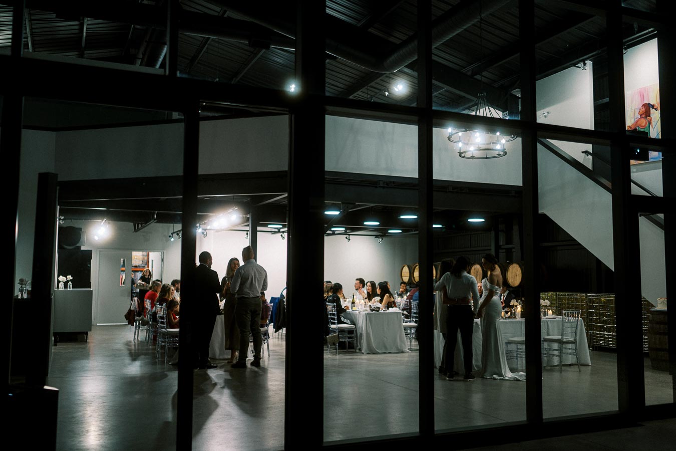 View of a modern indoor wedding reception, featuring elegantly dressed guests socializing, seated at white-clothed tables under contemporary lighting fixtures, in a stylish venue with a high ceiling and wine barrels in the background.